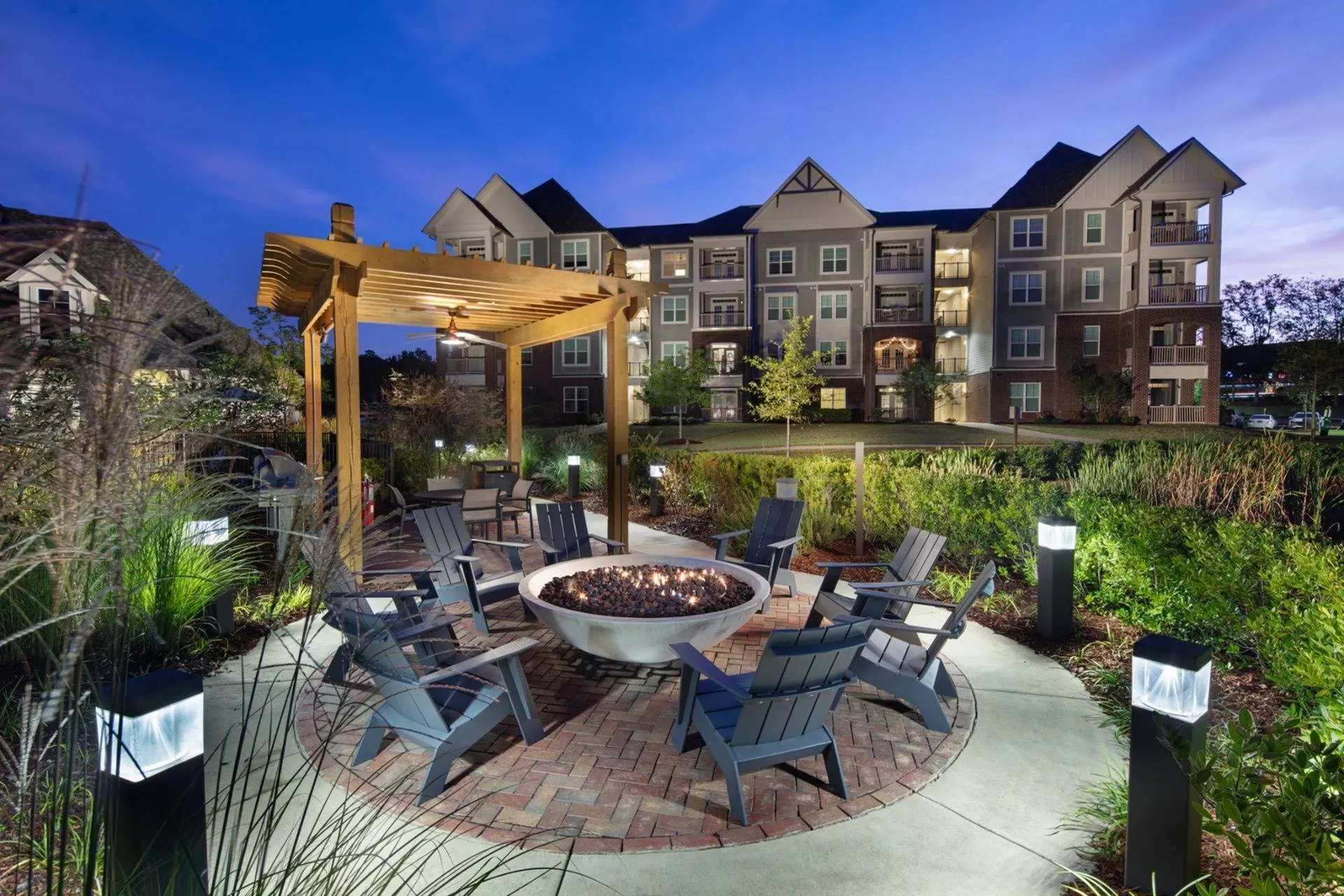 Vestavia Reserve A circular outdoor seating area with a fire pit and pergola, surrounded by greenery and lit lamp posts at dusk.