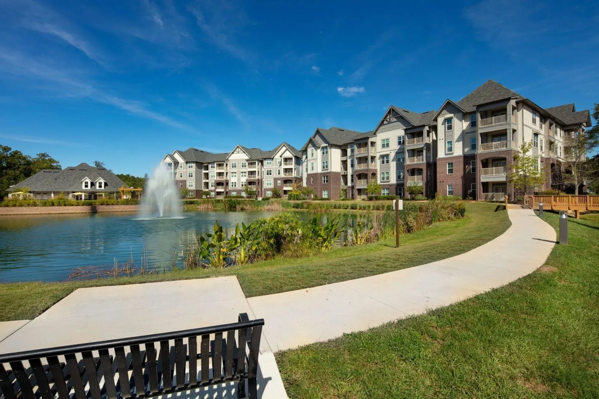 Vestavia Reserve A scenic view of apartment buildings with a pond, fountain, grassy area, and a bench under a clear blue sky.