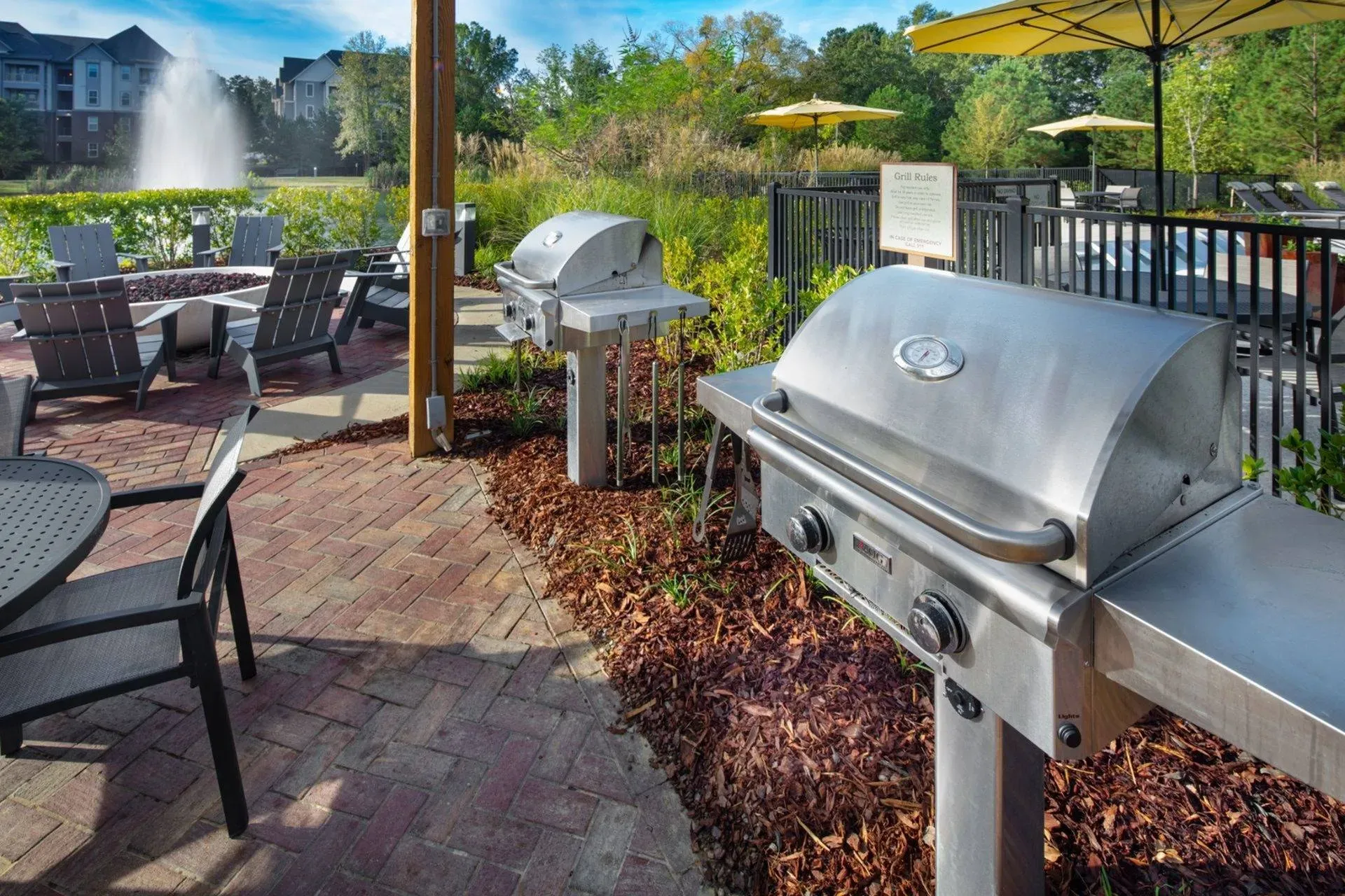 Vestavia Reserve Outdoor patio with two silver grills, tables, chairs, parasols, brick flooring, and a water fountain in the background.